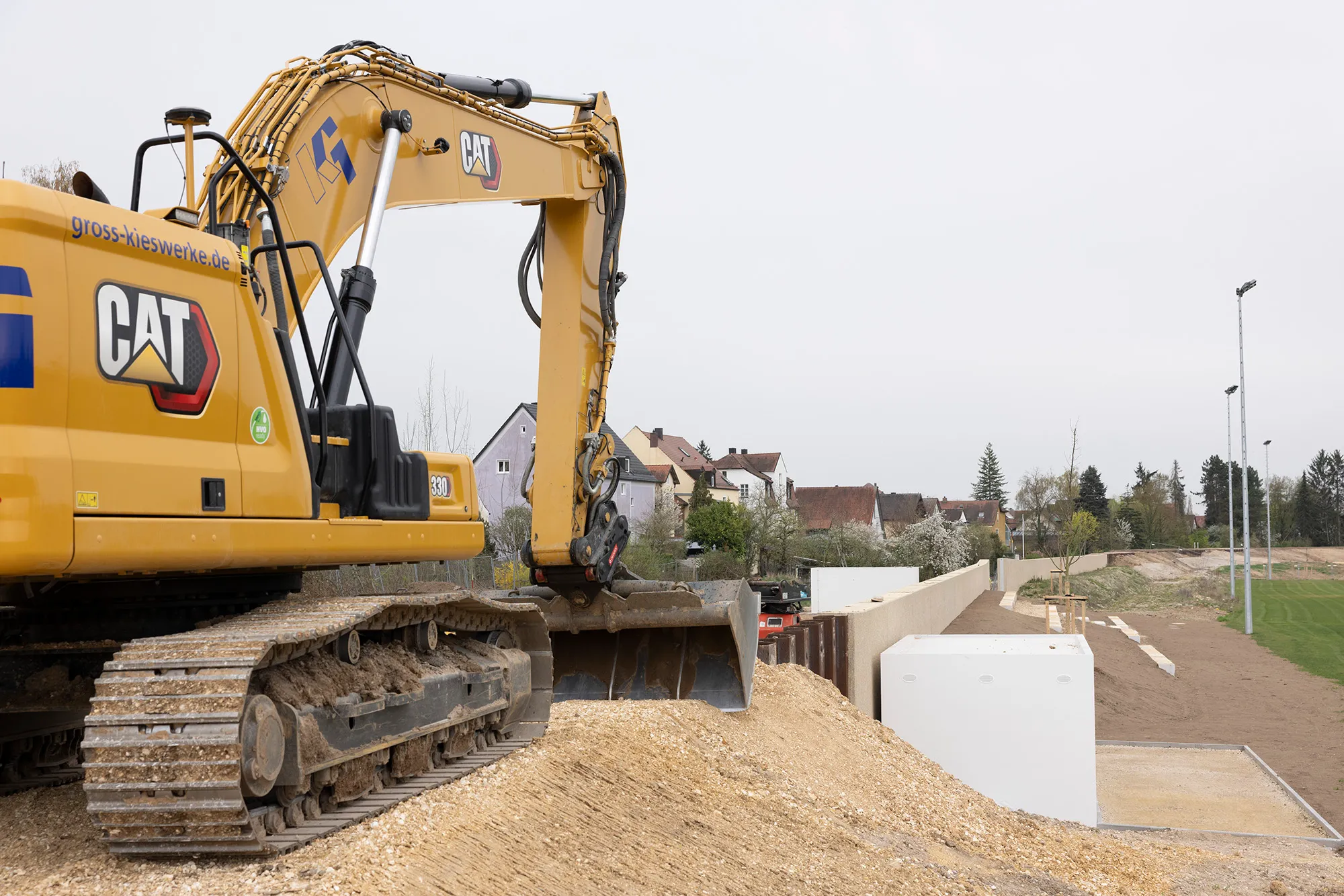 Gelber CAT-Kettenbagger auf Schotterhaufen neben einer fertiggestellten Hochwasserschutzmauer aus Beton; im Hintergrund Wohnhäuser am Ortsrand.