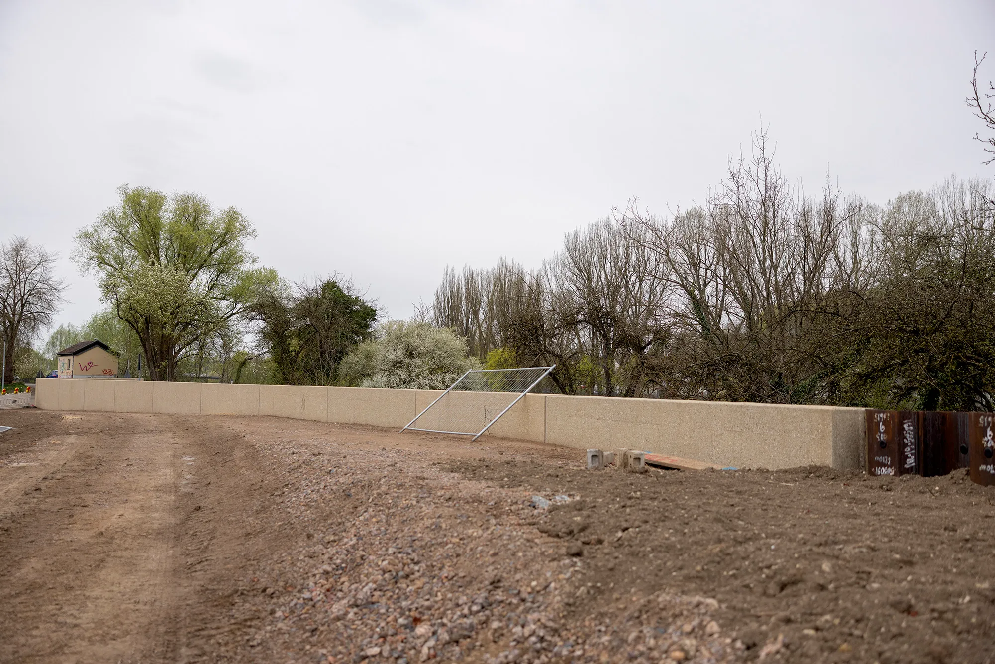 Lange, hüfthohe Hochwasserschutzmauer aus Beton mit Stahl-Schiebetor an der Donau; davor aufgewühlte Schotterfläche, dahinter kahle Bäume unter grauem Himmel.