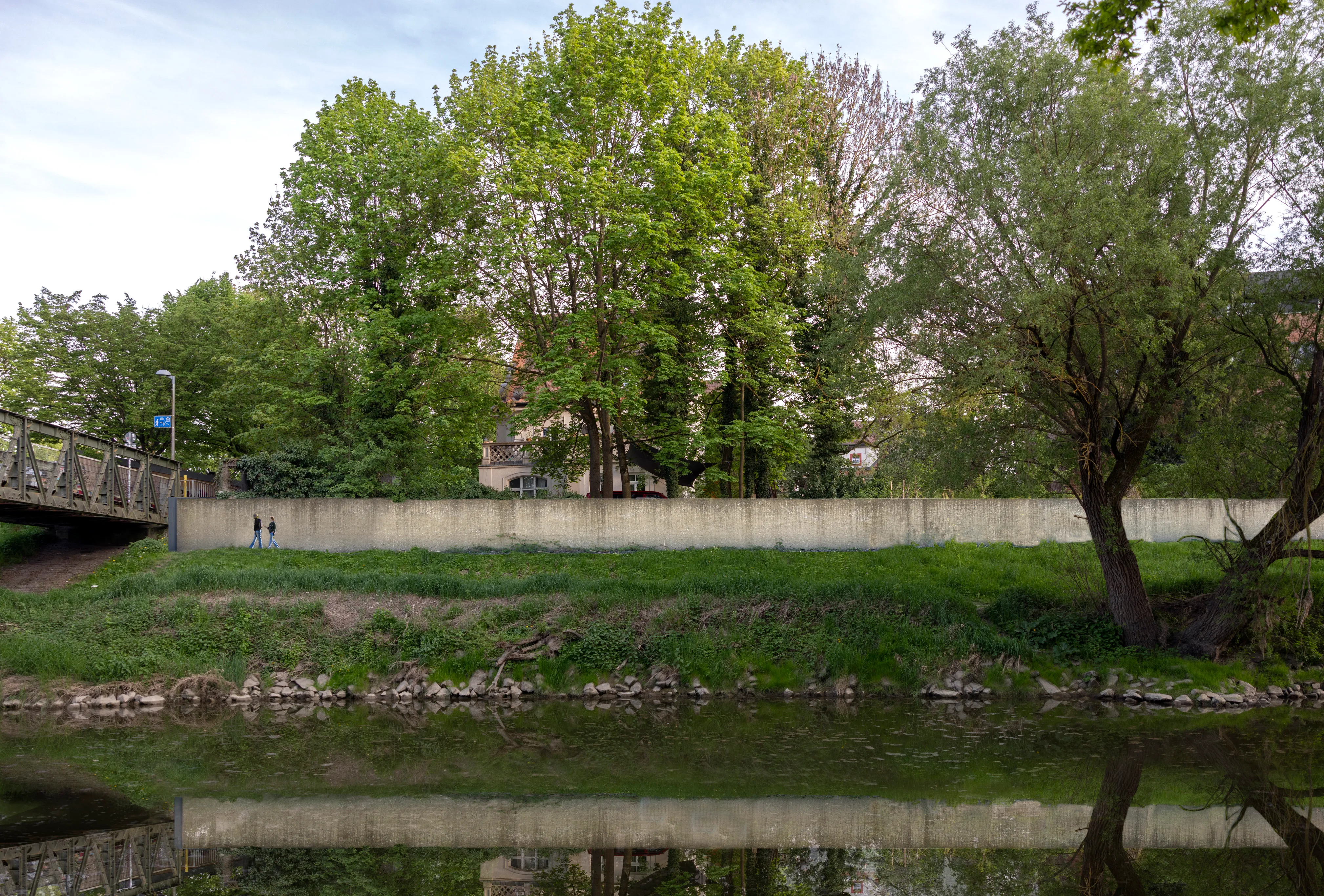 KI-Visualisierung: Lange Betonmauer entlang des Donauufers, die den Blick auf die dahinterliegenden Bäume und Häuser abriegelt; links eine kleine Brücke mit zwei Spaziergängern, im Vordergrund das ruhige Wasser mit Spiegelung unter frühlingsgrünen Bäumen.