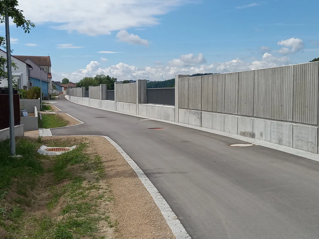 Asphaltierte Anliegerstraße, die rechts auf ganzer Länge von einer hohen Betonmauer begleitet wird; links Wohnhäuser am Ortsrand, im Hintergrund Hügel unter blauem Himmel mit Wolken.