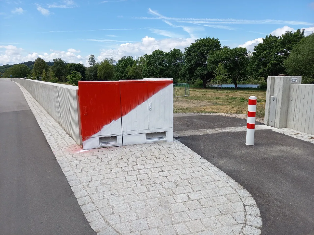 Fertige Hochwasserschutzmauer aus glattem Beton entlang eines gepflasterten Wegs; davor ein rot-weiß gestrichener Technikschrank und ein rot-weißer Sperrpoller, dahinter eine Wiese mit Bäumen unter blauem Himmel.