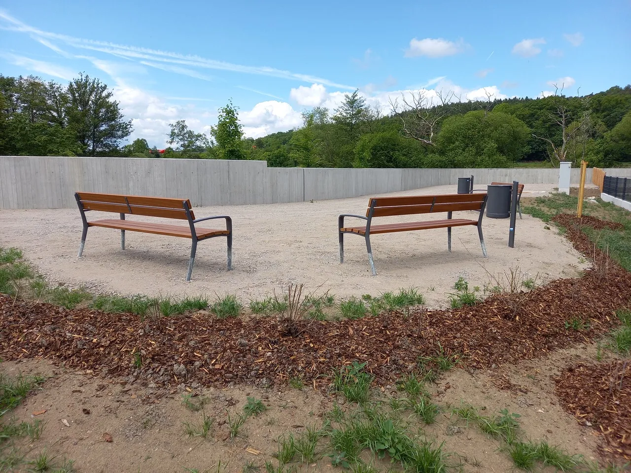 Zwei Holzbänke auf einem Schotterplatz vor einer durchgehenden grauen Betonmauer, die den Blick auf die dahinterliegende Landschaft abriegelt; im Vordergrund frisch angelegte Rindenmulch-Beete.